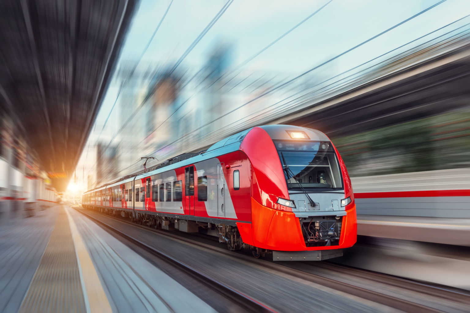 A modern red and gray passenger train speeds through a station, creating a motion blur effect. The platform is empty, and the sun is shining brightly in the background.
