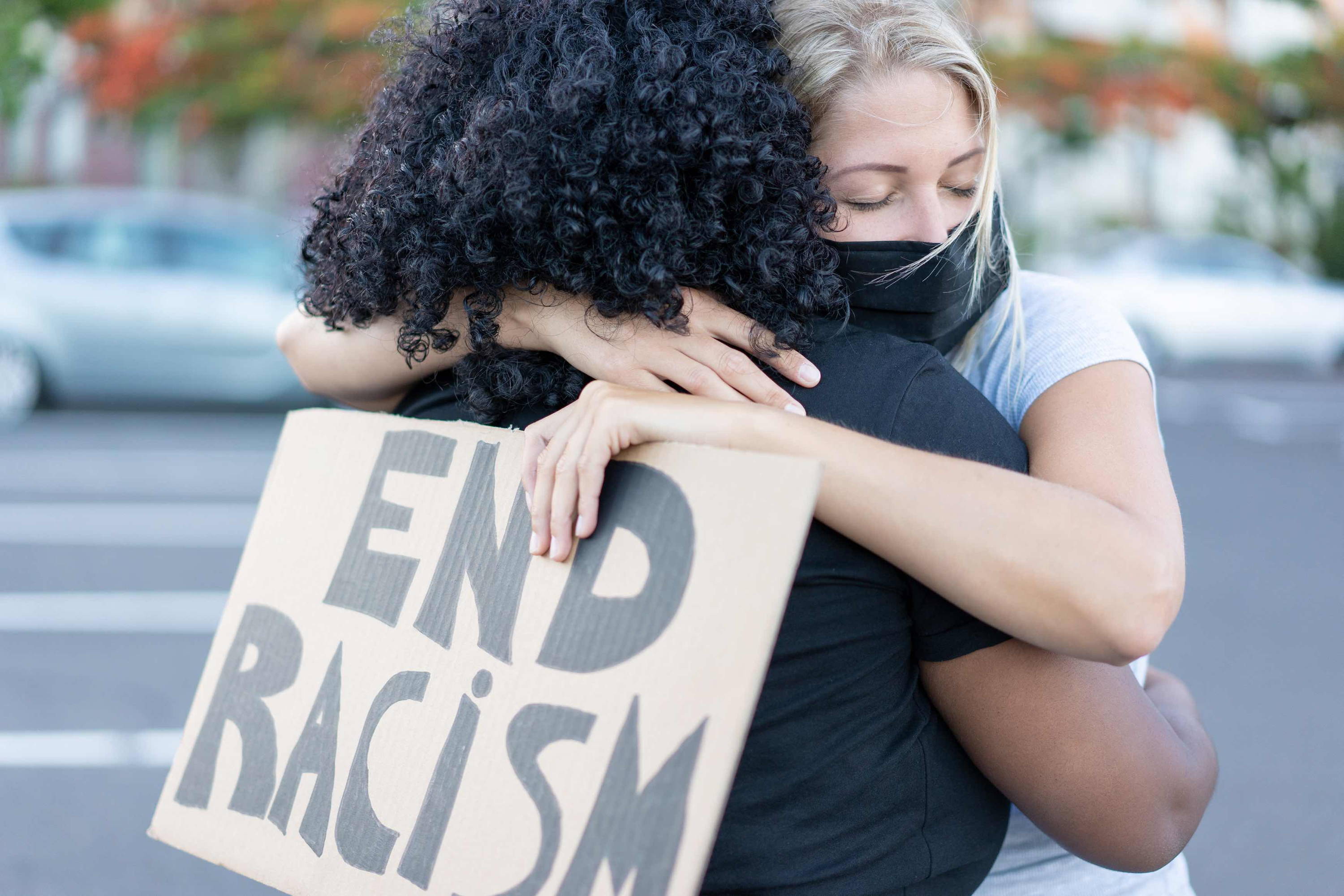 Black and white woman embrace holding End Racism placard