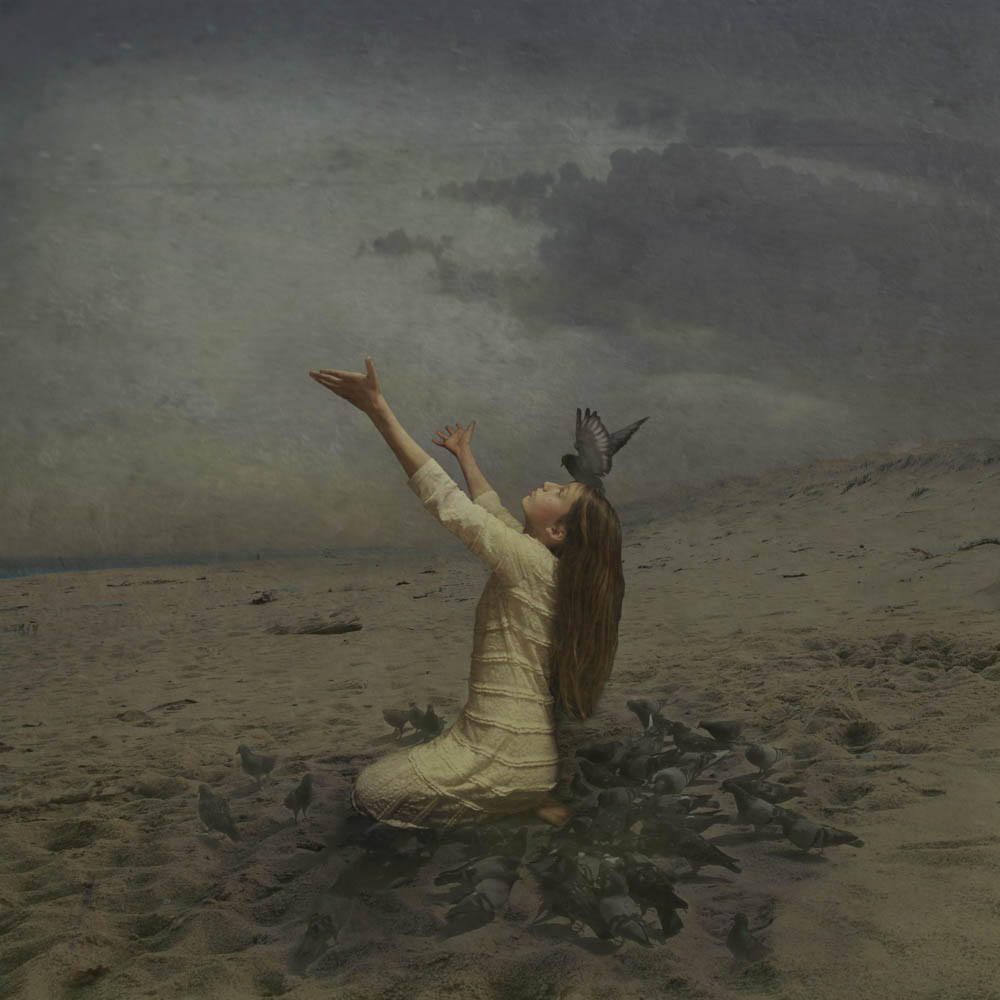 A girl sitting on the beach beneath a gray sky. Her arms are outstretched toward the sky and she looks up.