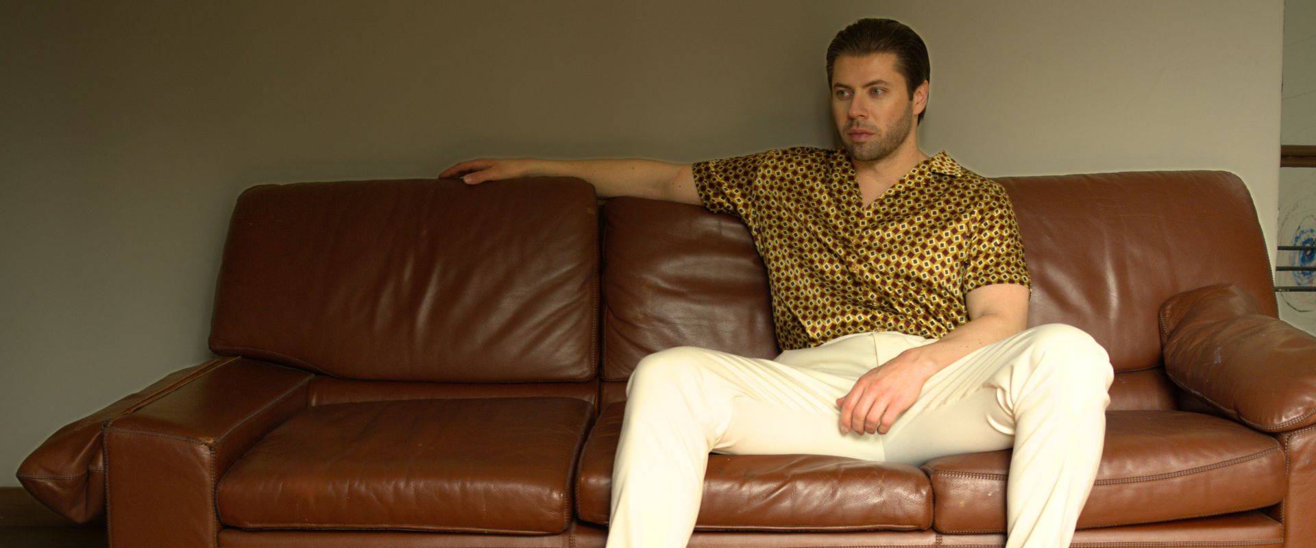 model sitting on a brown sofa wearing white trousers and a gold bowling silk shirt from 1000 kingdoms