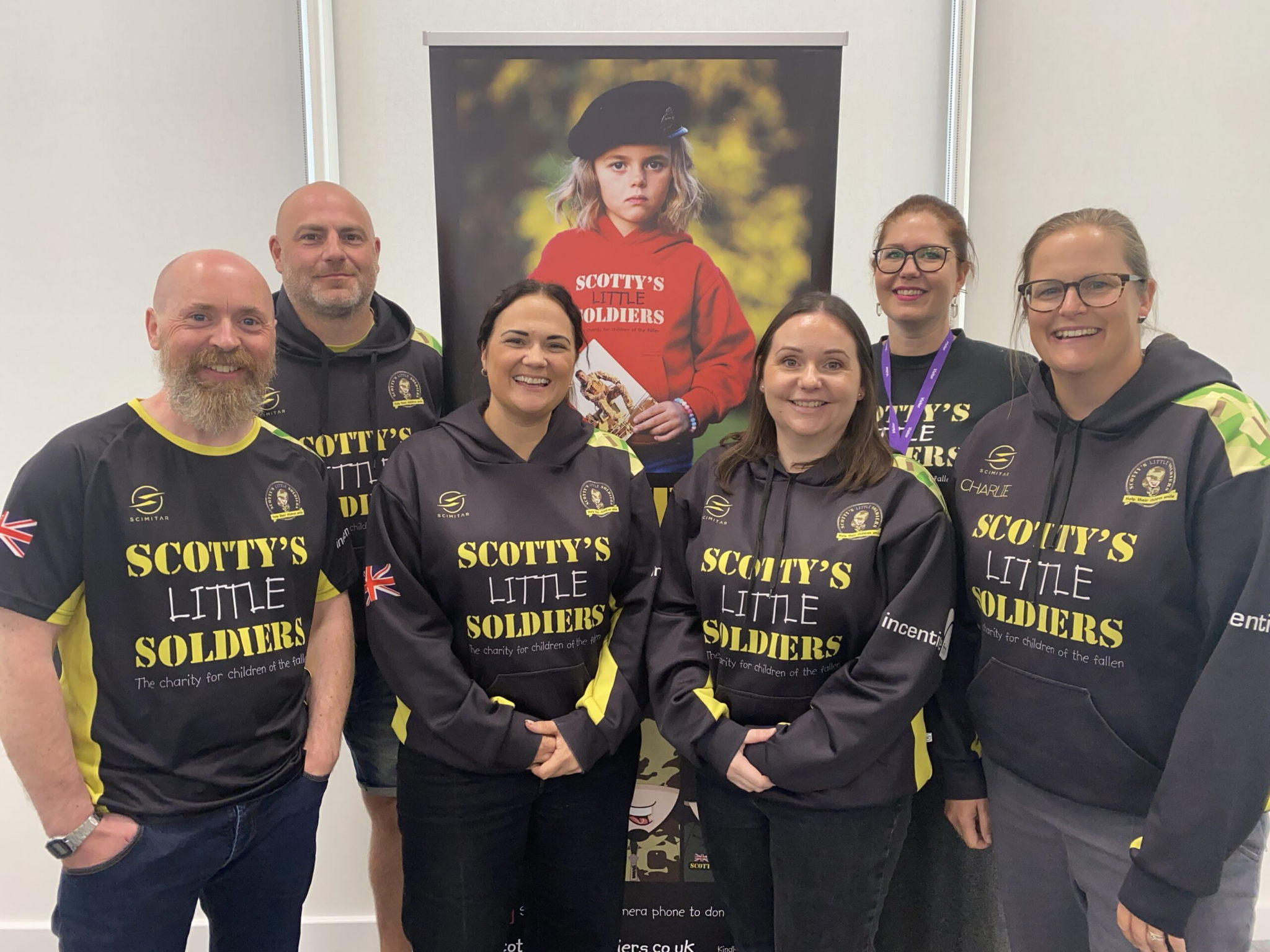 Six adults wearing matching Scottys Little Soldiers hoodies stand smiling in front of a banner showing a child in a red jacket and beret with the same logo.