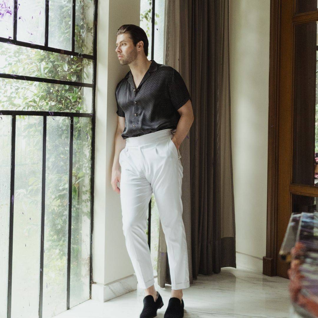 model leaning against a wall wearing black velvet shoes white high rise pants and a short sleeve blacked dotted silk shirt from 1000 Kingdoms