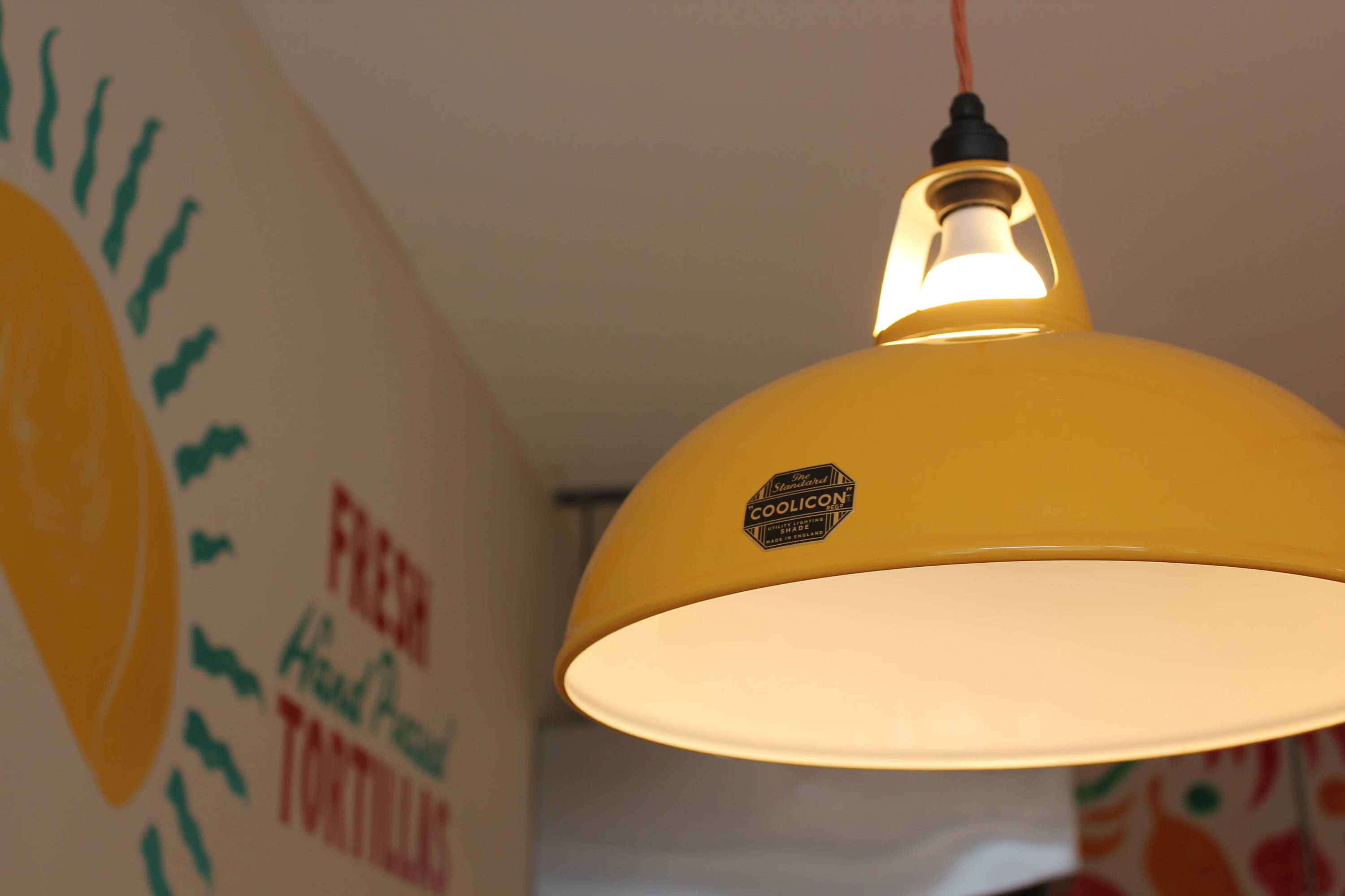 A close-up of a large Yellow Lampshade with an LED bulb and Black Lampholder with red cable. The Coolicon badge is cleary in focus. In the background is a handpainted sign on the wall.