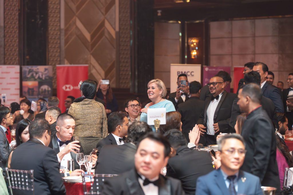A group of people in formal attire are seated at round tables during a gala or award event. Some are standing and smiling, while others are applauding. The atmosphere appears lively and celebratory.