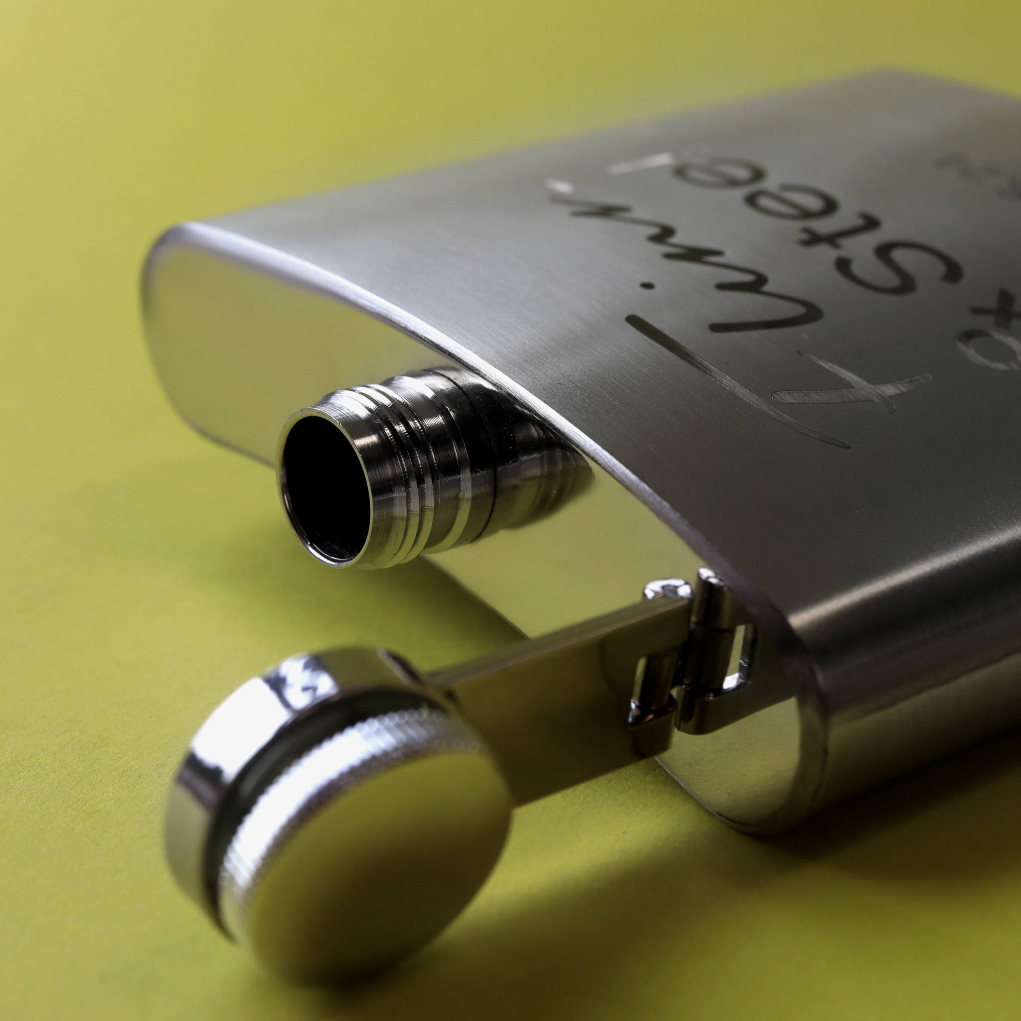 A close up image of a stainless steel hip flask on a lime green background. The flask is laid down with the top open and a Flinn & Steel logo engraving on the side.