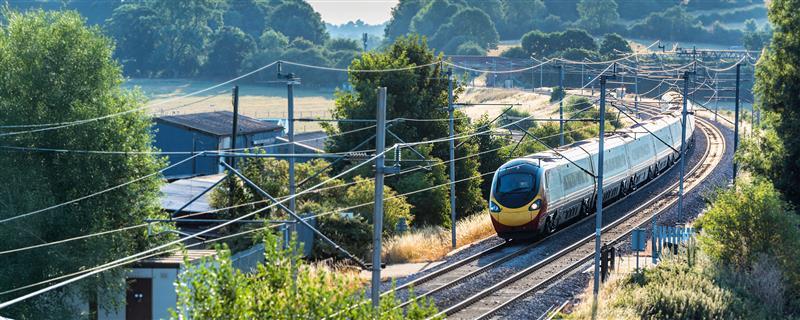 A modern passenger train travels along curved, electrified tracks through a rural landscape with trees, fields, and overhead power lines on a sunny day.