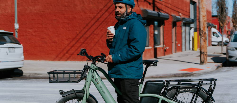 a man drinking holding a coffee while standing over an electric cargo bike