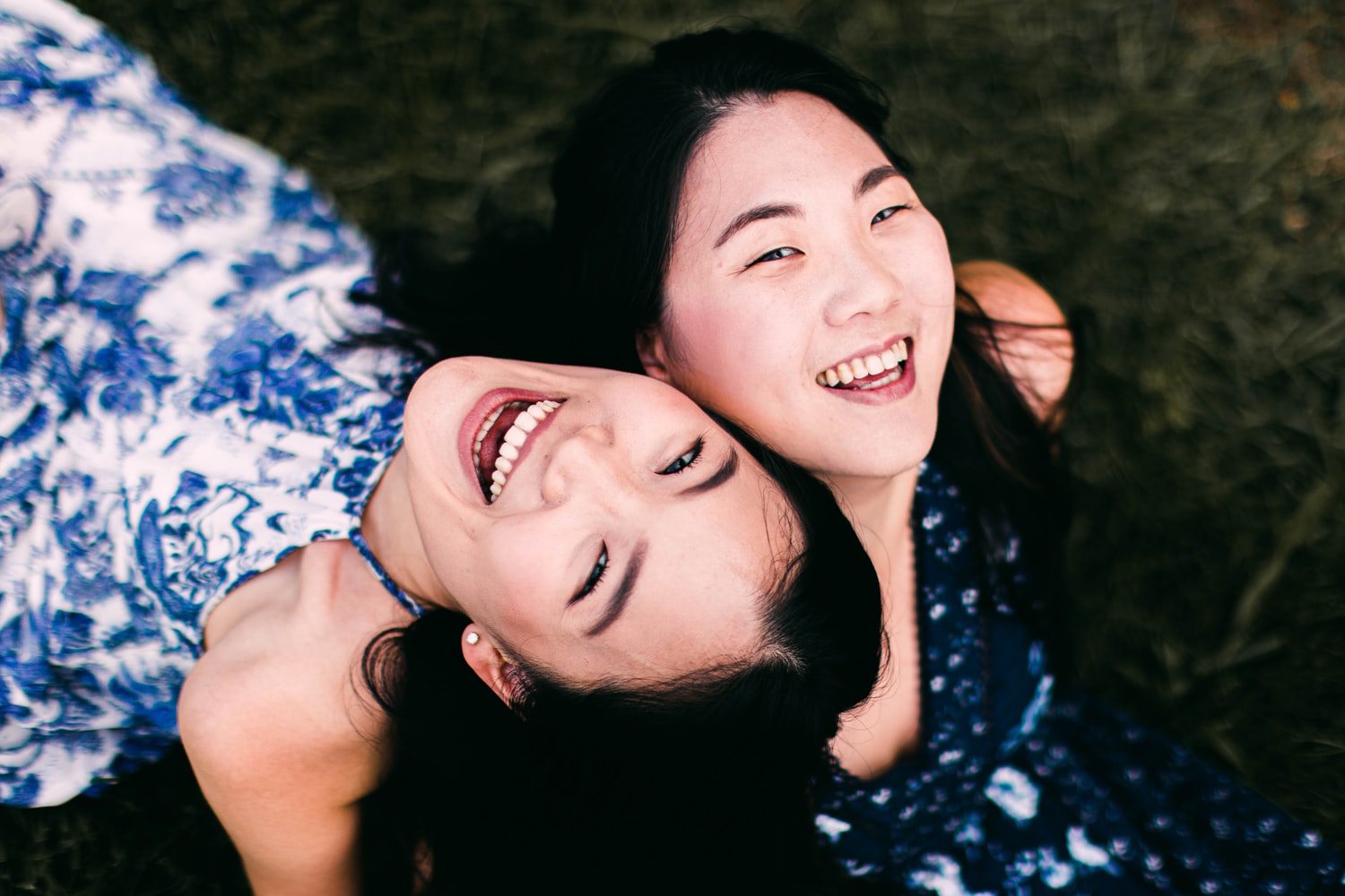 Two attractive asian women laugh and sit back to back looking to the camera.