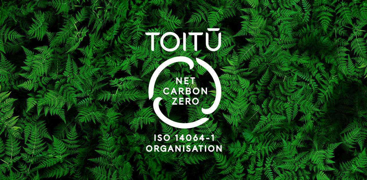 White Toitū net carbon zero ISO 14064-1 organisation logo over a background of dense green fern leaves, symbolizing environmental sustainability and carbon neutrality.