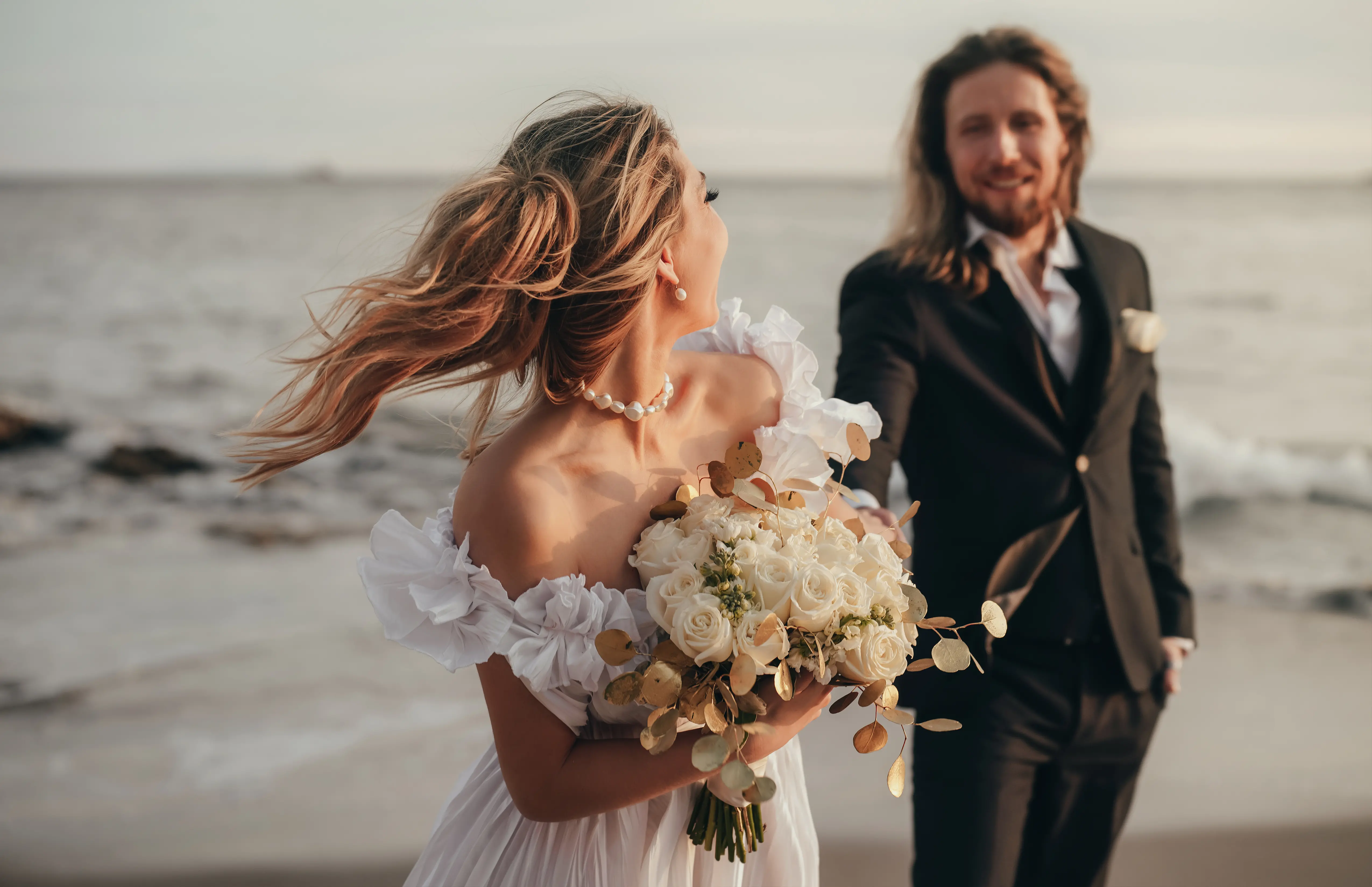 Une belle mariée blonde aux longs cheveux se tient dans une robe de mariée bohème blanche en arrière-plan des rochers.