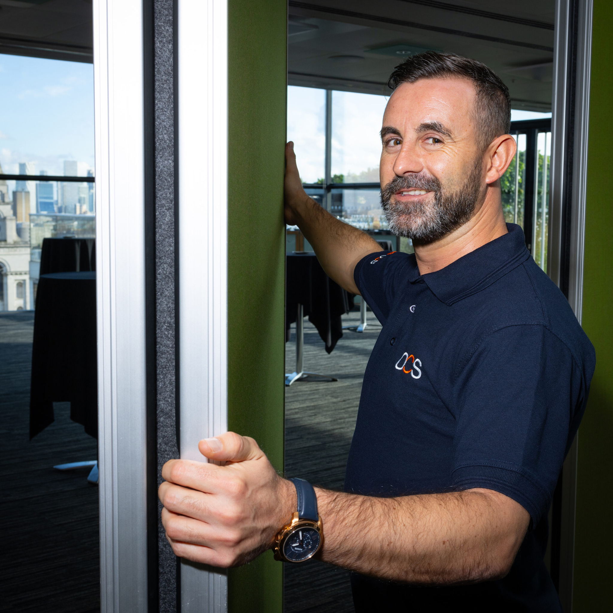 A man with a beard is smiling at the camera while opening a sliding panel in a modern room. He is wearing a dark polo shirt with a small logo and a watch on his wrist. A city skyline is visible through the large windows in the background.