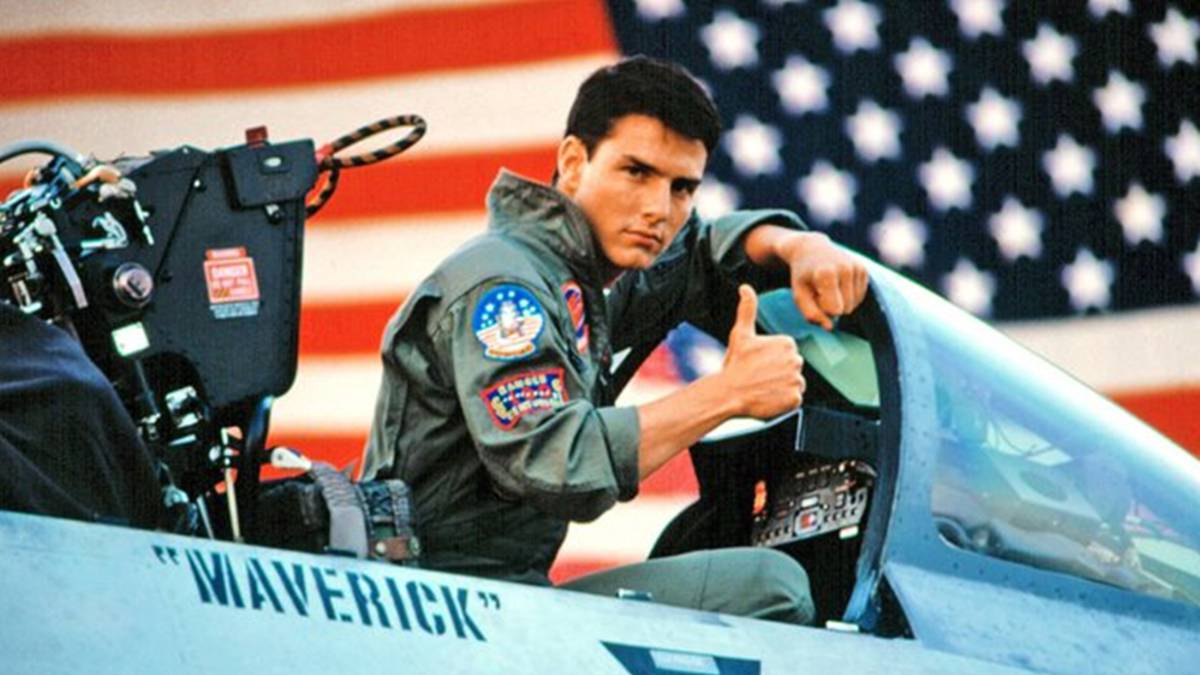 Maverick giving the thumbs up sitting inside a one of his fighter planes with a large american flag behind him.