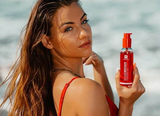 Olje Shine Brown Watermelon Oil, ki ga drži zagorela rjavolaska ob morju.