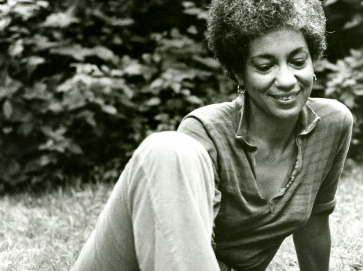 A young June Jordan sitting in the grass wearing a loose shirt smiling looking down.