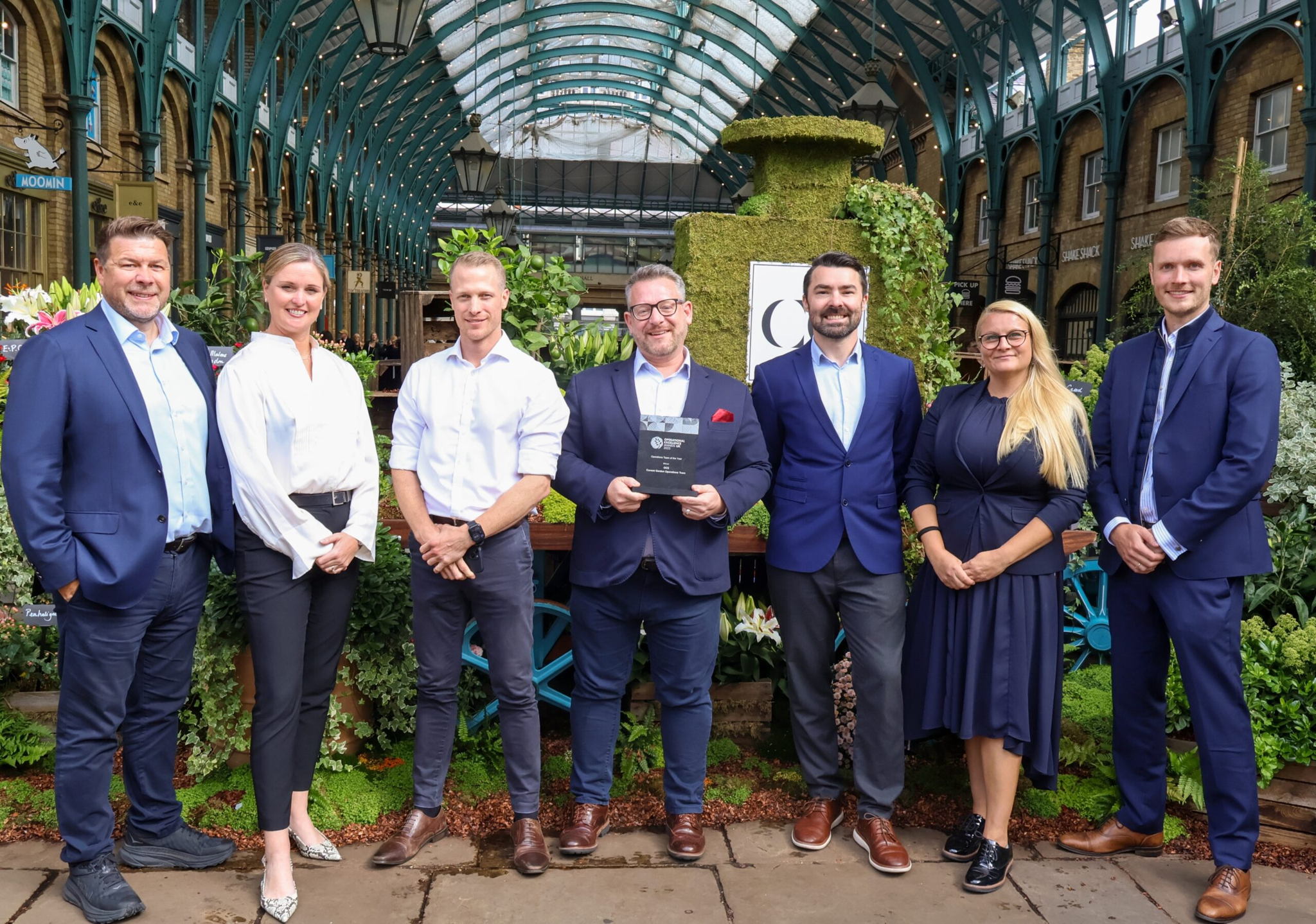 Seven people in business attire stand in a row outdoors under a glass-roofed structure, smiling at the camera. The group is surrounded by lush greenery and flowers, and one person holds a plaque or award.
