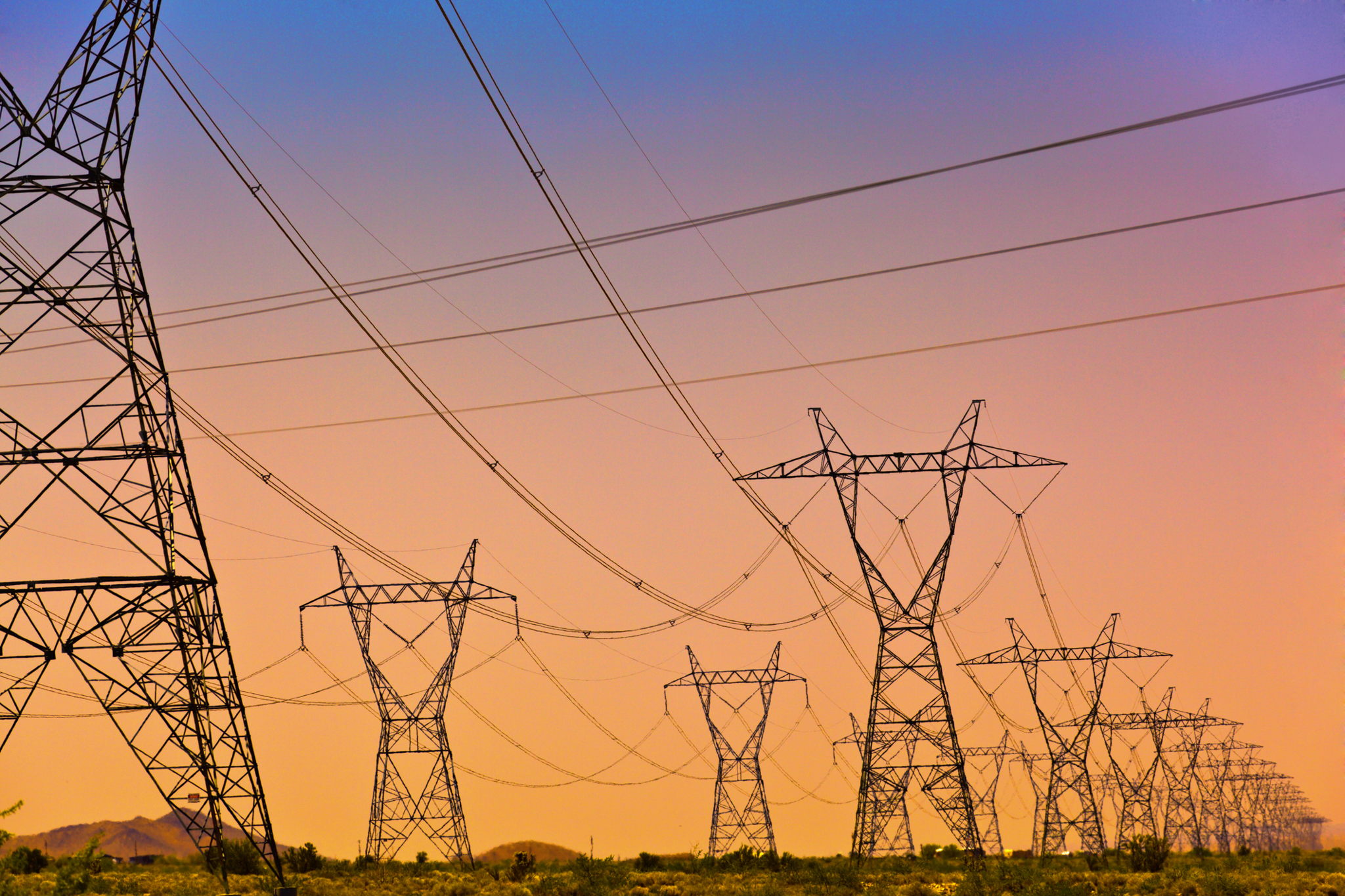 Multiple high-voltage power lines and transmission towers stretch across a landscape at sunset, with a colorful sky transitioning from blue to orange and low hills in the background.