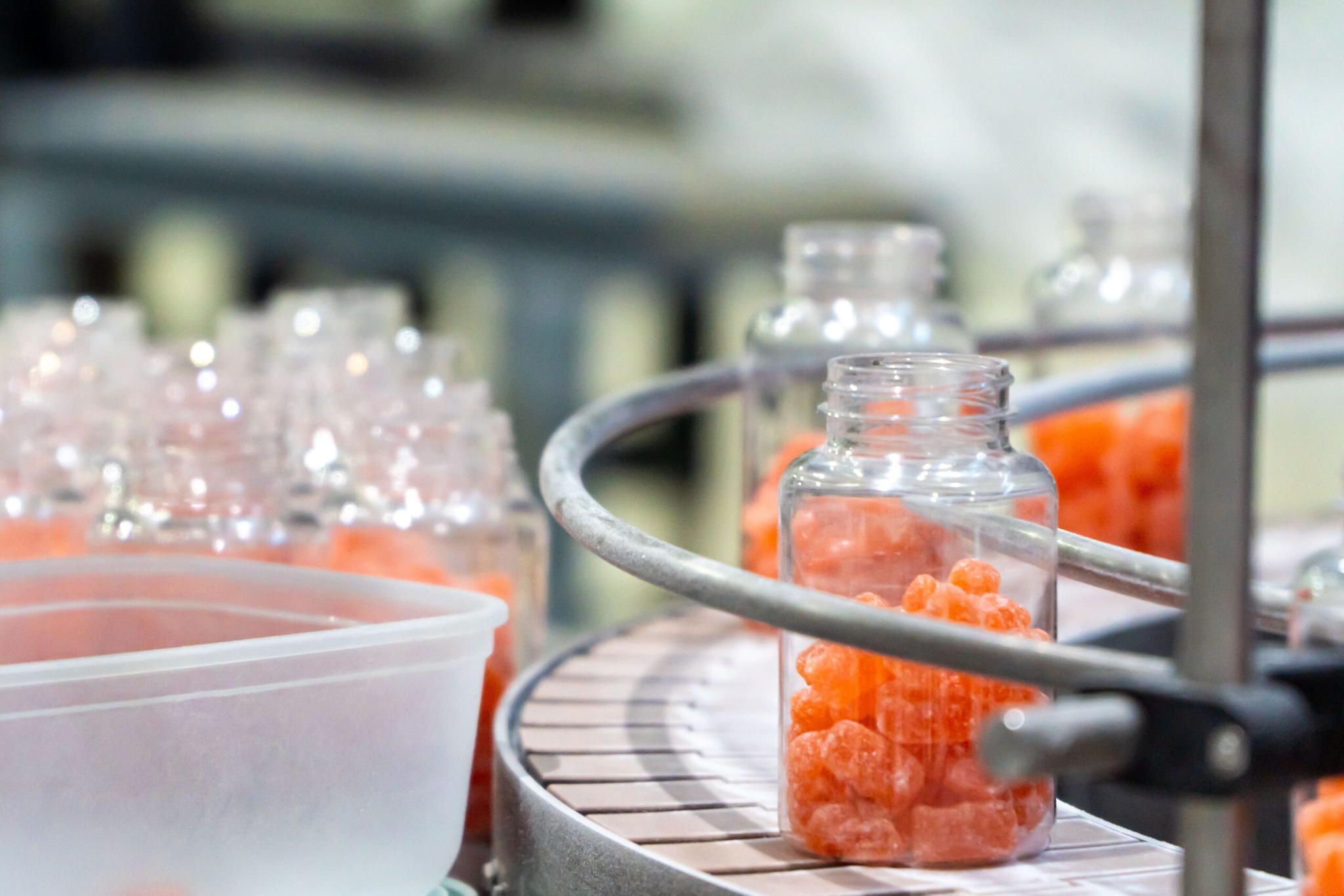 Clear plastic bottles being filled with orange gummy vitamins on a conveyer belt in a factory setting, with a container and more bottles in the background.