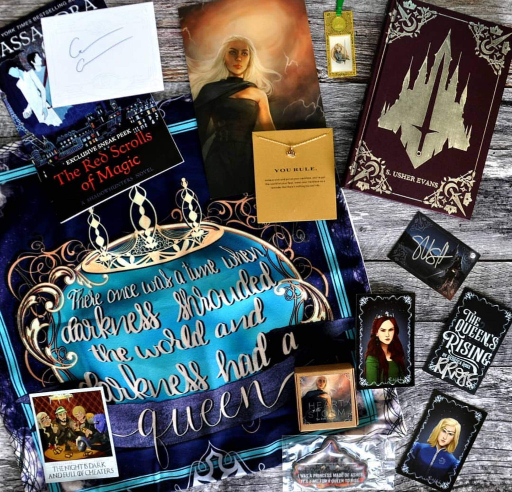 April 2019 Hold My Crown box theme included The Red Scrolls of Magic Sampler, Collector's Polaroid inspired by Game of Thrones, Queen's Rising/Resistance Character Cards, Ash Princess Air Freshener, ROAR inspired Soap, Metal Bookmark, Crown Necklace, Young Elites inspired Pillowcase, City of Veils by S. Usher Evans, and Three Dark Crowns Shirt for Seelie and Solitary Fae boxes only. 