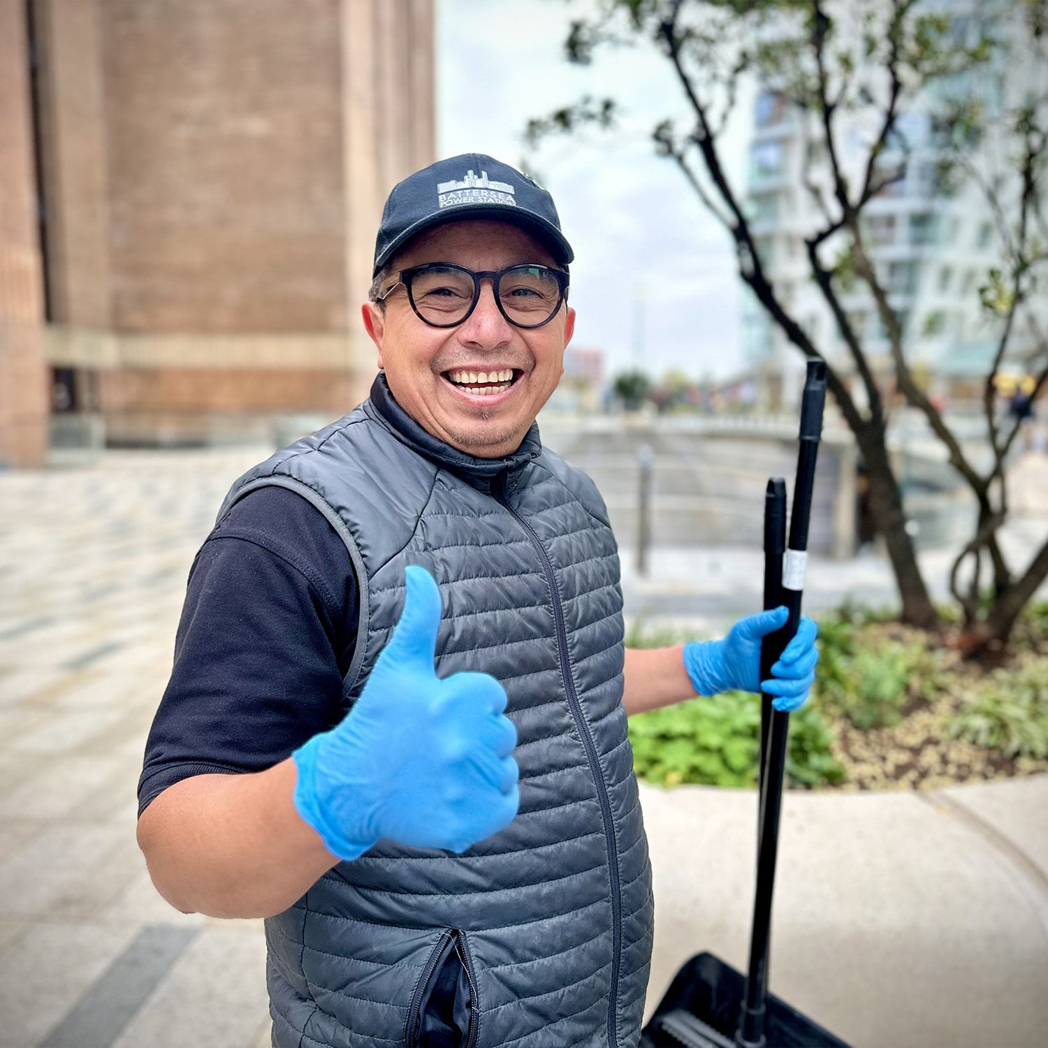 A smiling person wearing glasses, a black cap, a gray vest, and blue gloves gives a thumbs-up while holding a broom outside near modern buildings and trees.