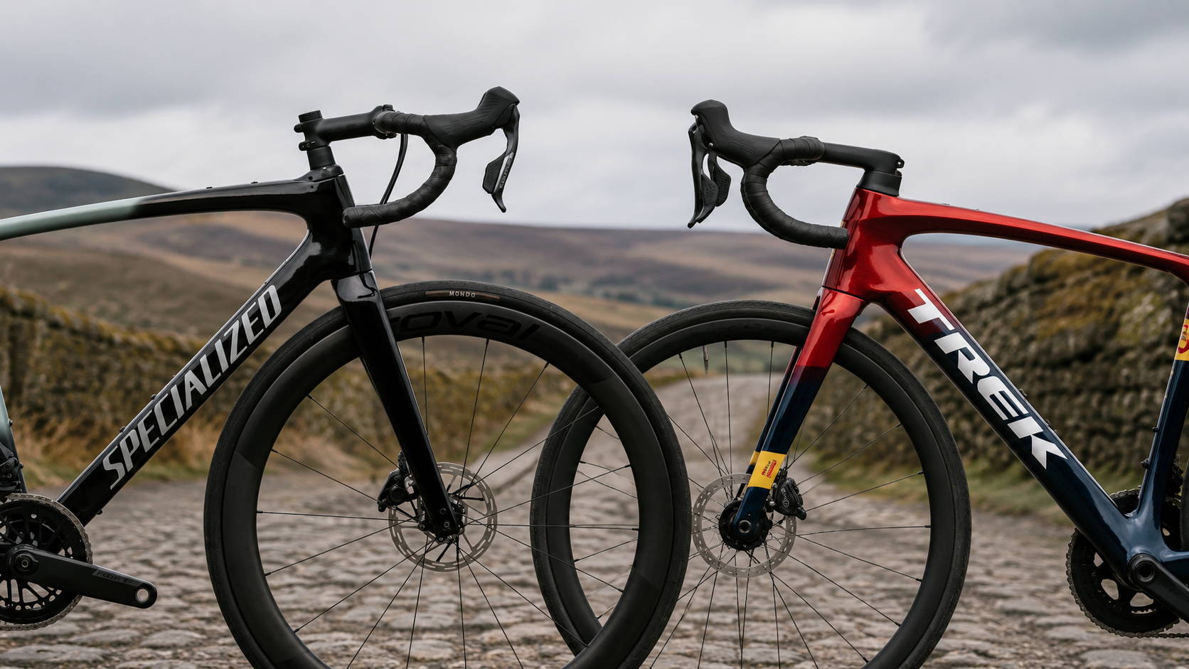 Specialized Roubaix en Trek Domane staan frontaal tegenover elkaar op een ruwe kasseiweg, ingezoomd op de sturen, voorvorken en voorwielen als visuele vergelijking tussen twee endurance-racefietsen.