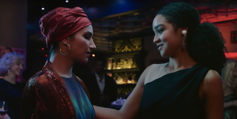 Kat reaching out to Adena who is wearing a headscarf, both are in a bar with dim lights.