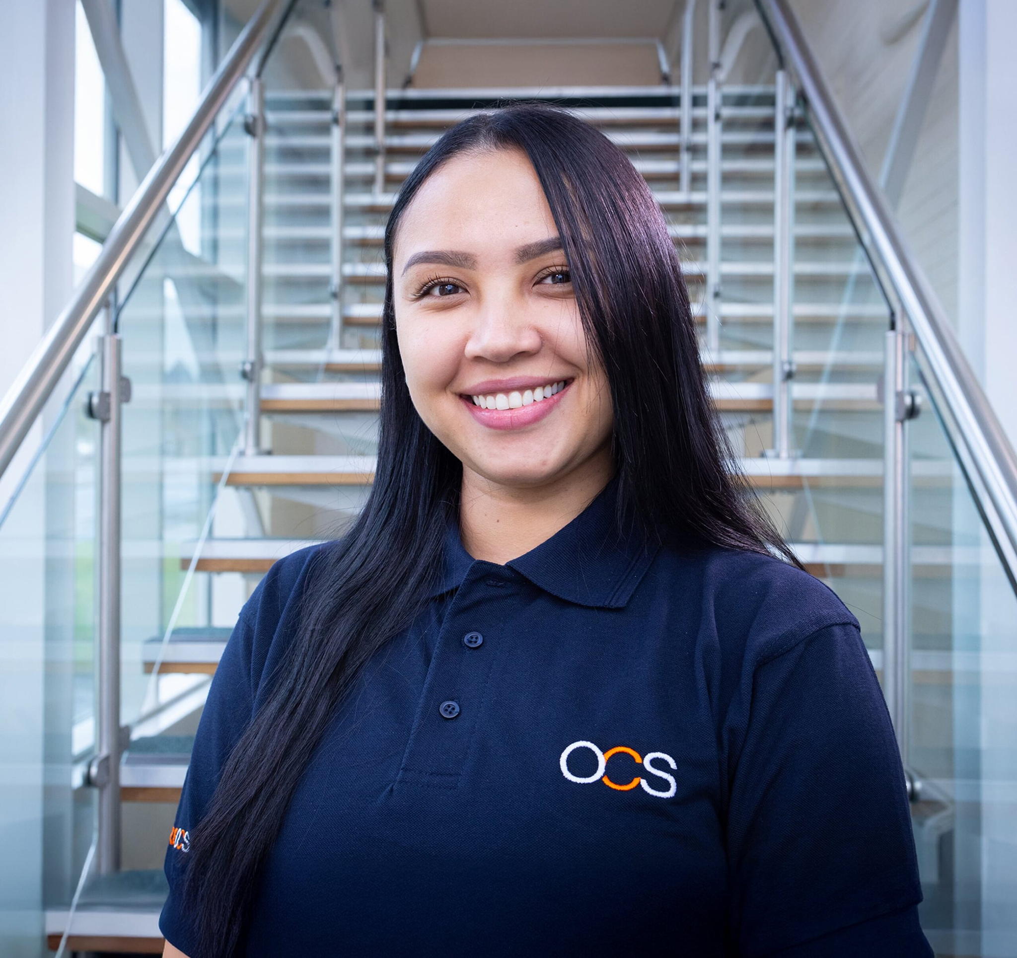 A woman with long dark hair stands smiling in front of a staircase, wearing a navy blue polo shirt with the OCS logo on it. The setting appears bright with large windows in the background.