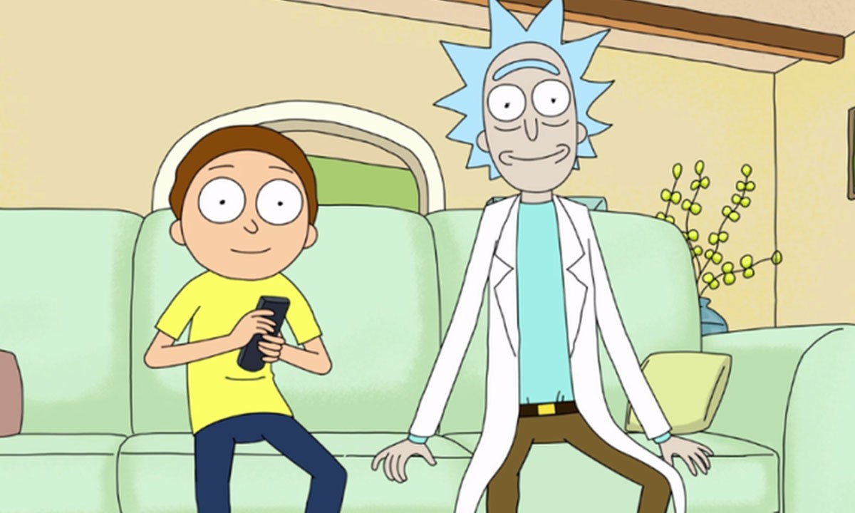 Photo of Rick and Morty sitting in their couch smiling staring at the camera with Morty holding the control.