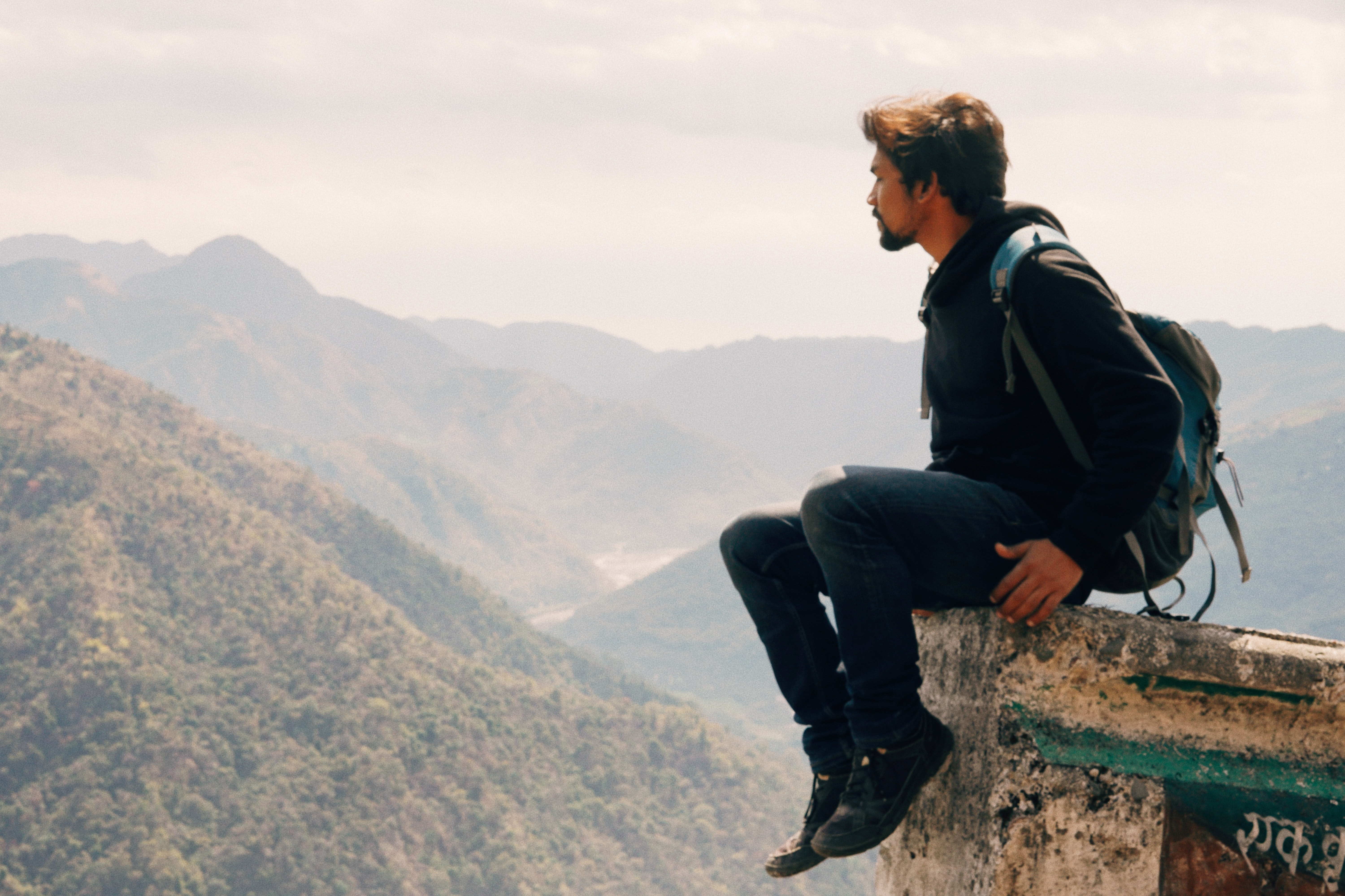 A man sits on the edge of a small cliff overlooking large mountains. His face is turned.