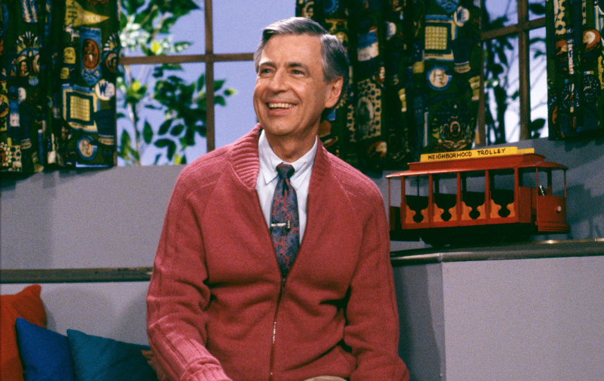 Mr. Rogers with his classic zip up sweater, sitting on a couch smiling looking at someone off camera.