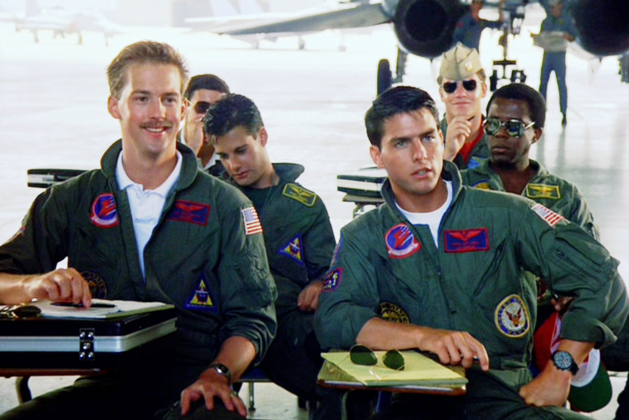 Maverick and Goose sitting in desks with others in their uniform, listening to a speaker. Goose is smiling and Maverick has a serious look.