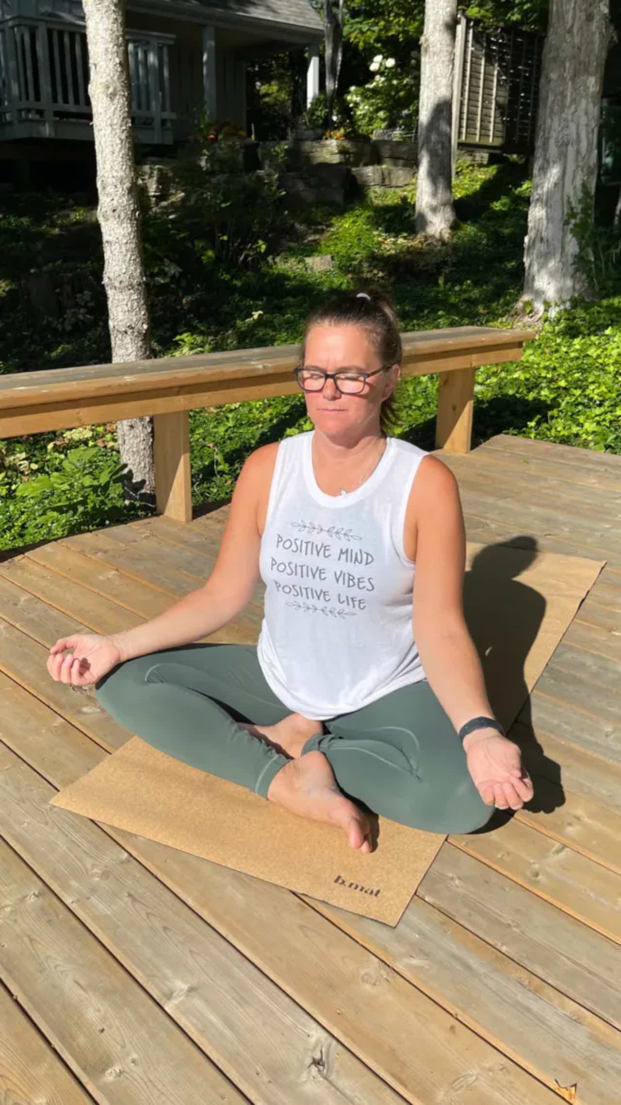 Woman sits cross-legged on a wooden deck in yoga pose, meditating with eyes closed, outdoors.