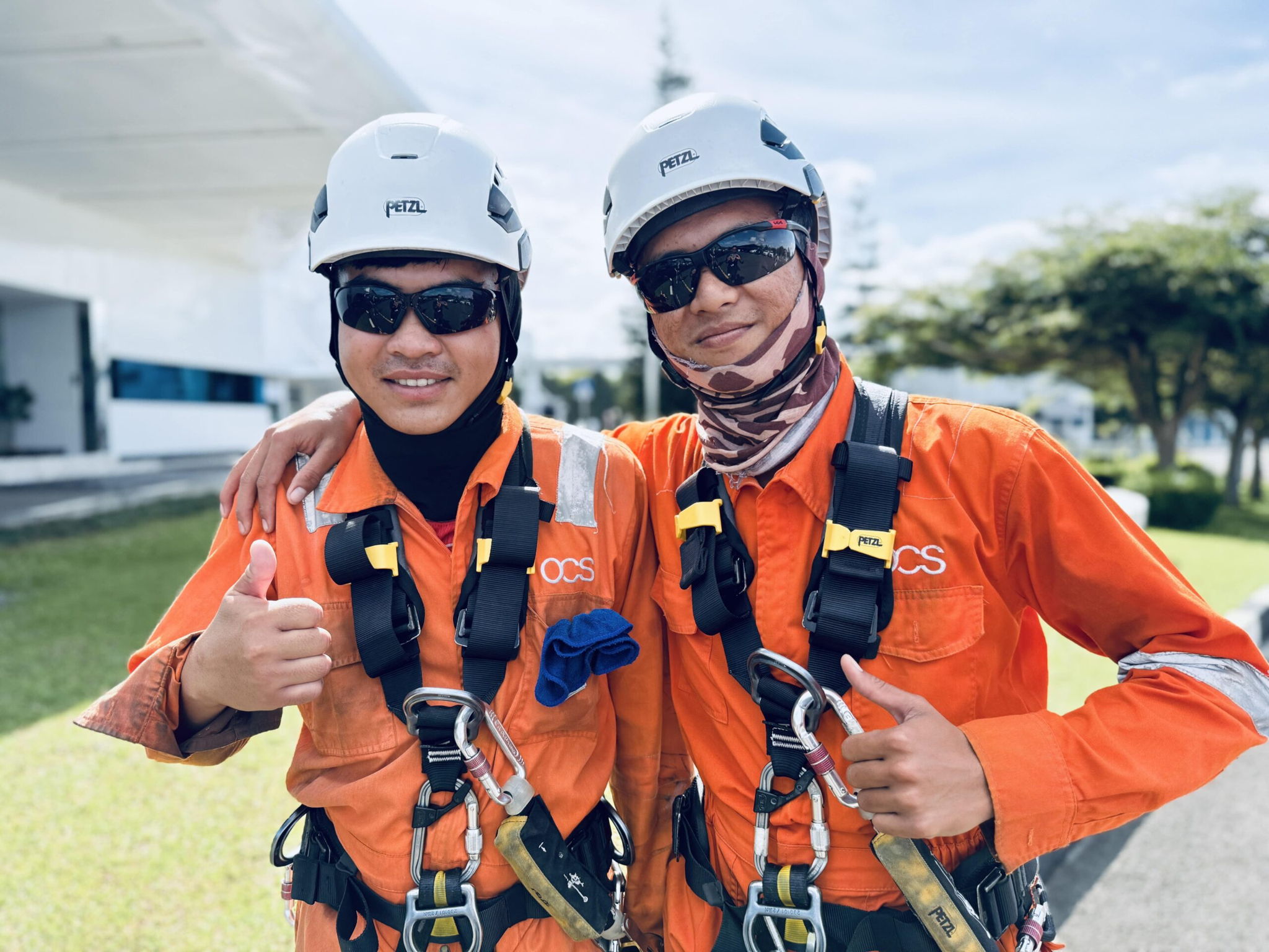 Two workers in orange safety uniforms, harnesses, helmets, and sunglasses smile and pose with thumbs up outdoors on a sunny day.