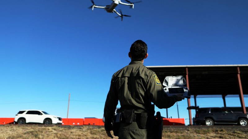 A man in a green uniform and utility belt looks up at a drone against a blue sky