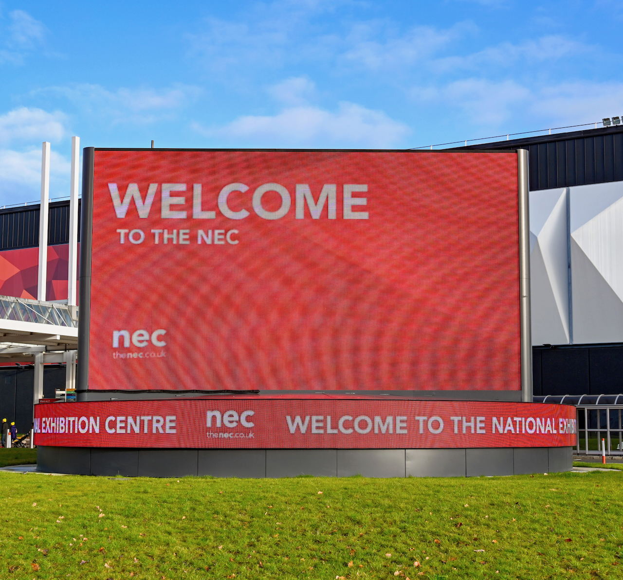 A large digital screen outside displays “Welcome to the NEC” at the National Exhibition Centre, with clear blue sky and green grass in the foreground.