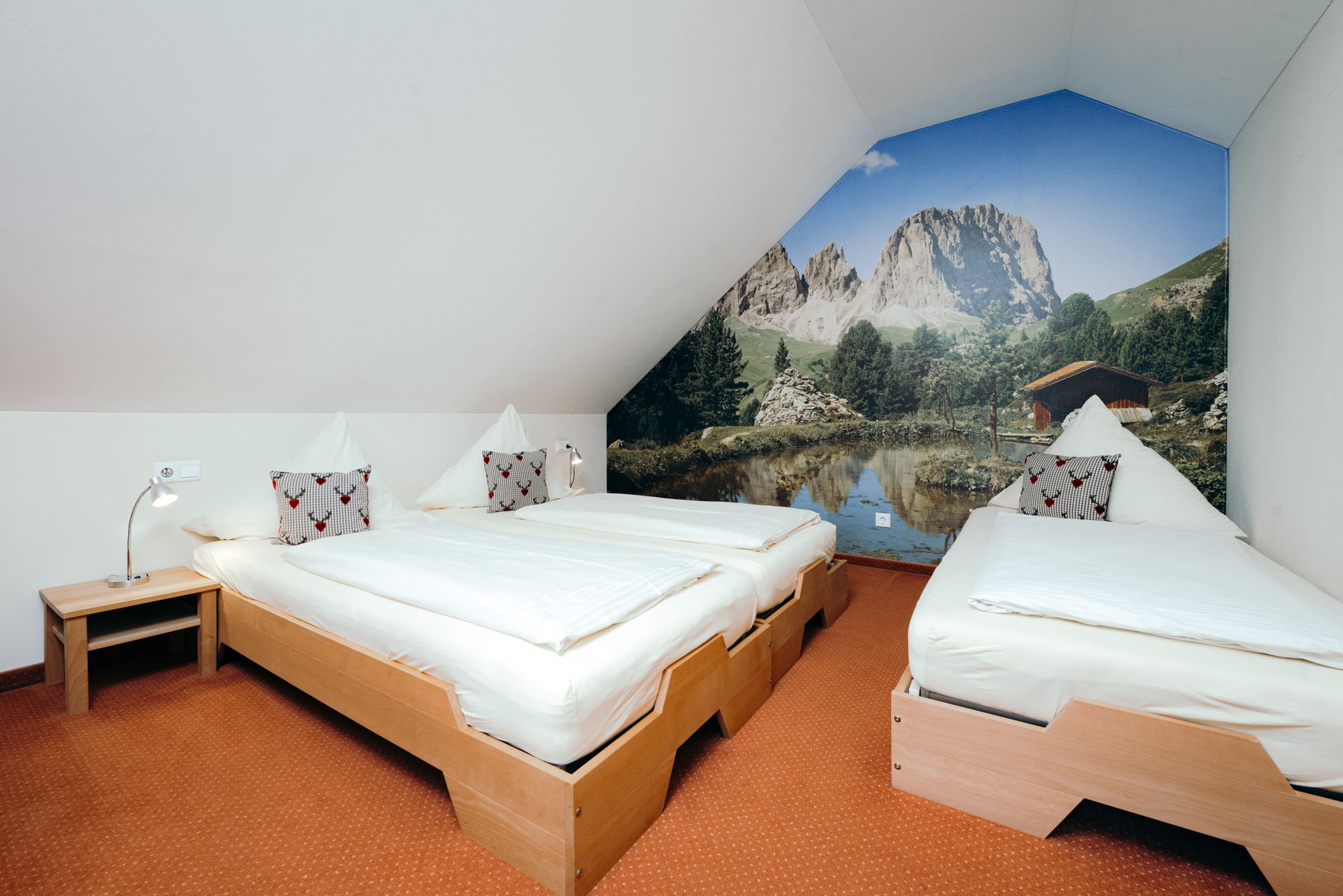 Ein Schlafzimmer mit Dachschräge und drei weiß bezogenen Einzelbetten, an der Wand ein buntes Bild von Bergen und Natur.
