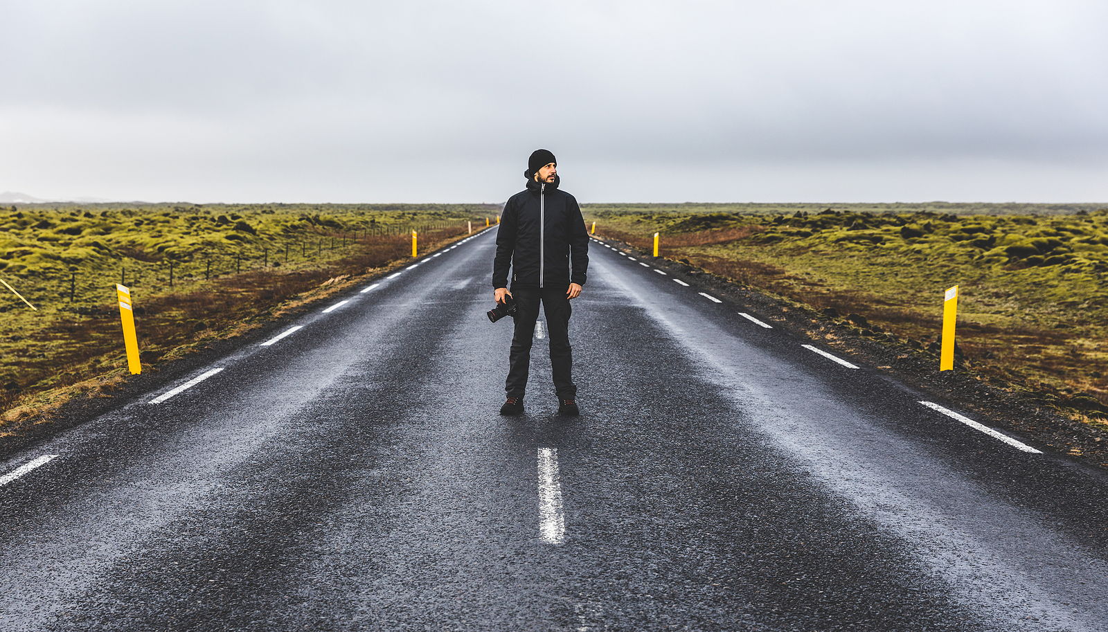 Young man wearing warm clothes standing in the middle of the Ring Road in Iceland looking away from camera.