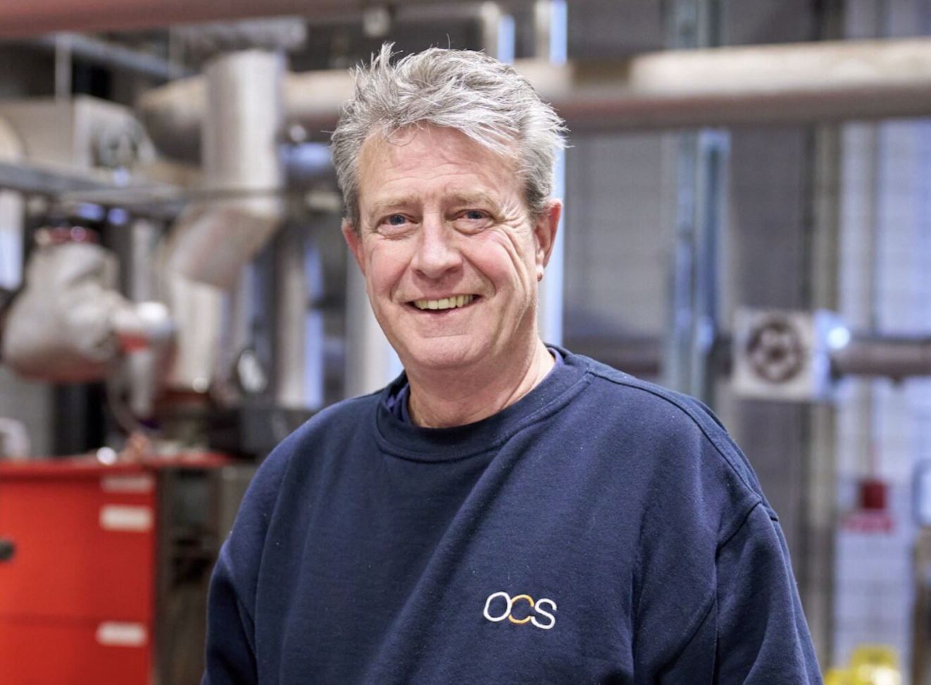 A smiling older man with gray hair wearing a navy blue sweatshirt with OCS on it stands indoors, with industrial pipes and equipment in the background.