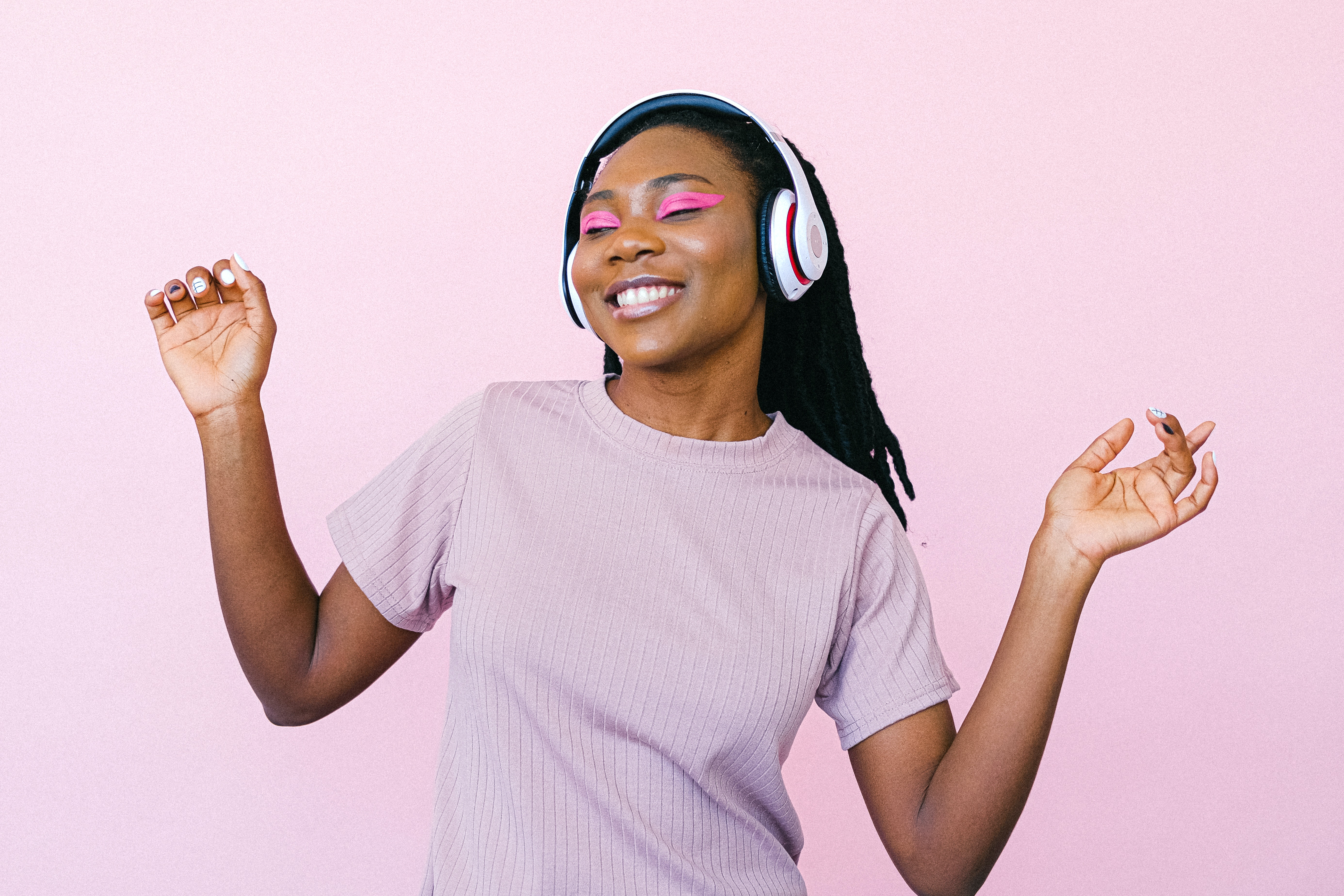 Image of black woman with headphones on and her eyes closed, dancing by herself. There is a purple/pink background.