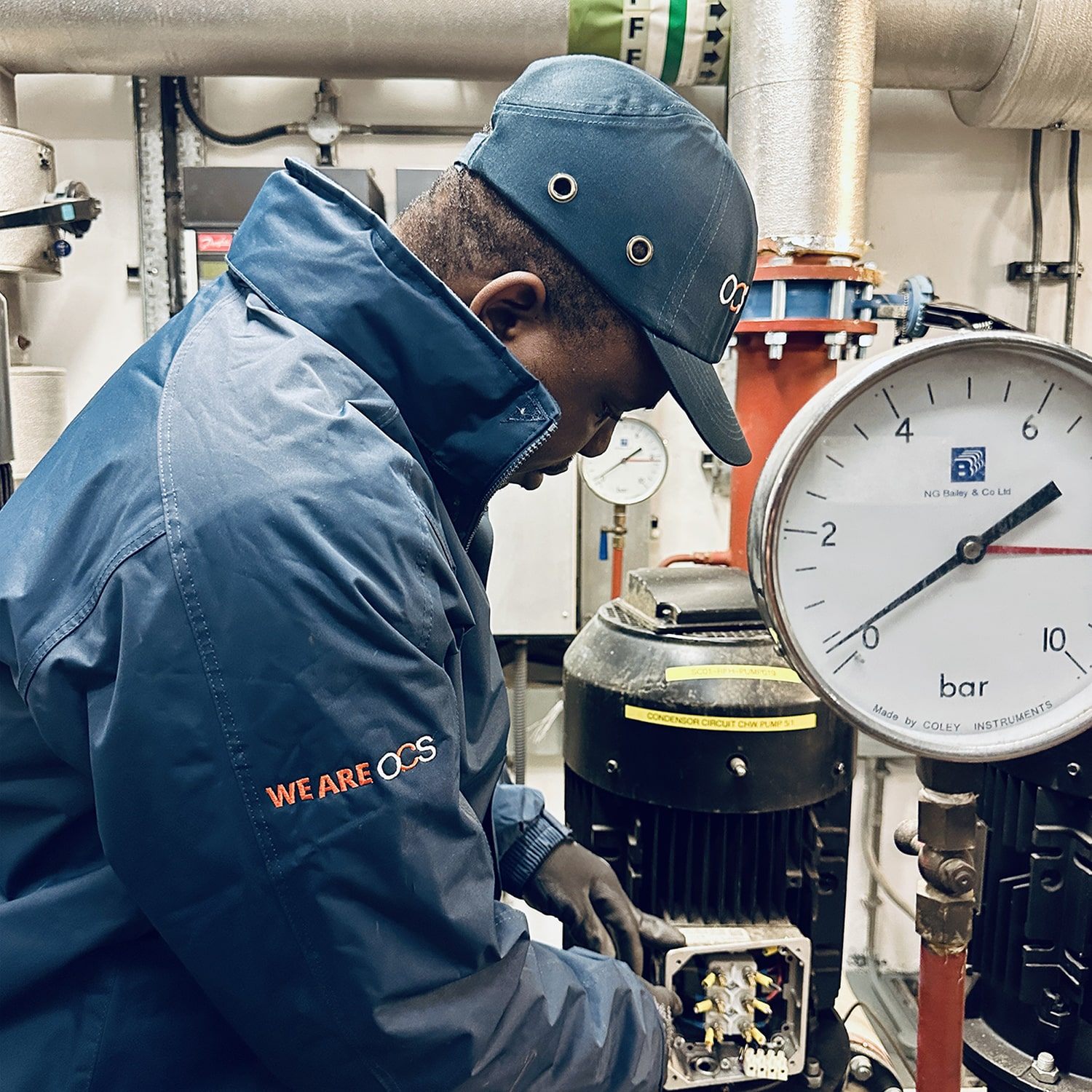 A technician in a blue jacket and cap labeled WE ARE OCS adjusts equipment near a large pressure gauge inside an industrial facility. Pipes and machinery surround the workspace.