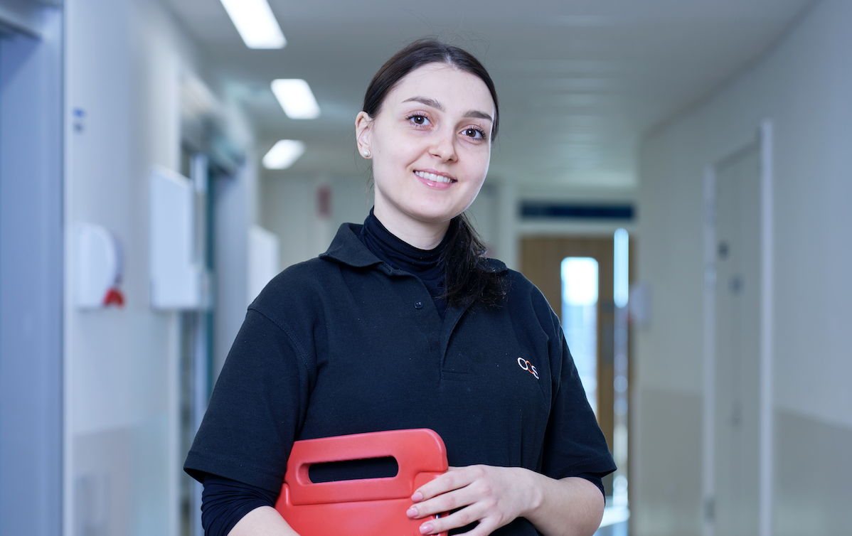 A person wearing a black polo shirt with a logo stands in a well-lit hallway, holding a red folder. The hallway has white walls, doors, and overhead lights. The person is smiling and looking forward.