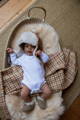 A baby laying on their back, on top of a neutral gingham blanket on a long haired sheepskin in a basket. the baby has a sheepskin bobble hat 