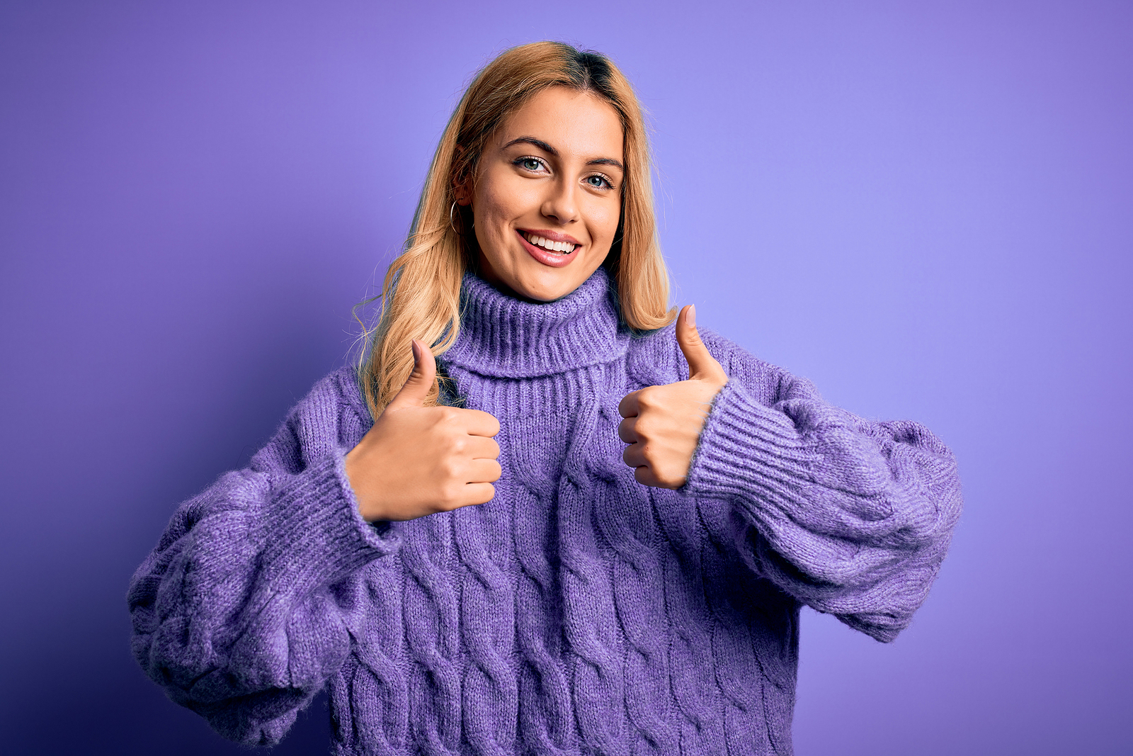 A blond woman with a large sweater smiles and holds her hands in a thumbs up gesture