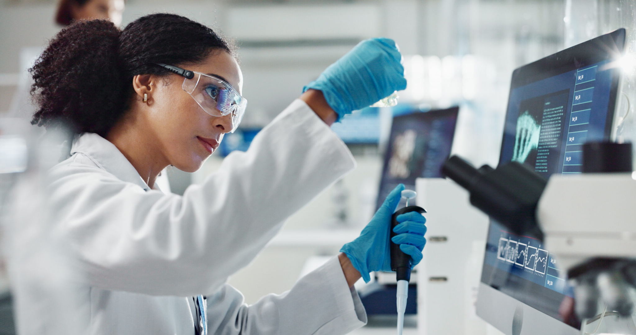 A scientist wearing safety goggles and gloves uses a pipette while working in a modern laboratory, with a computer monitor displaying data and scientific equipment visible on the desk.