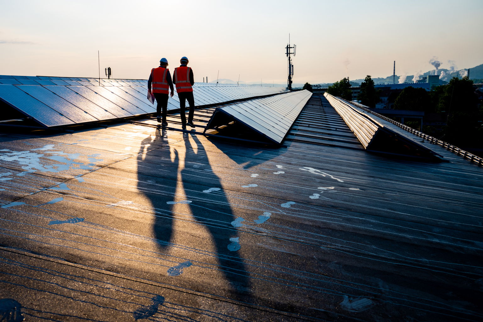 Two people wearing hard hats and orange safety vests walk on a roof covered with solar panels. The sun casts long shadows of them on the roof. The background includes industrial buildings and a hazy sky.
