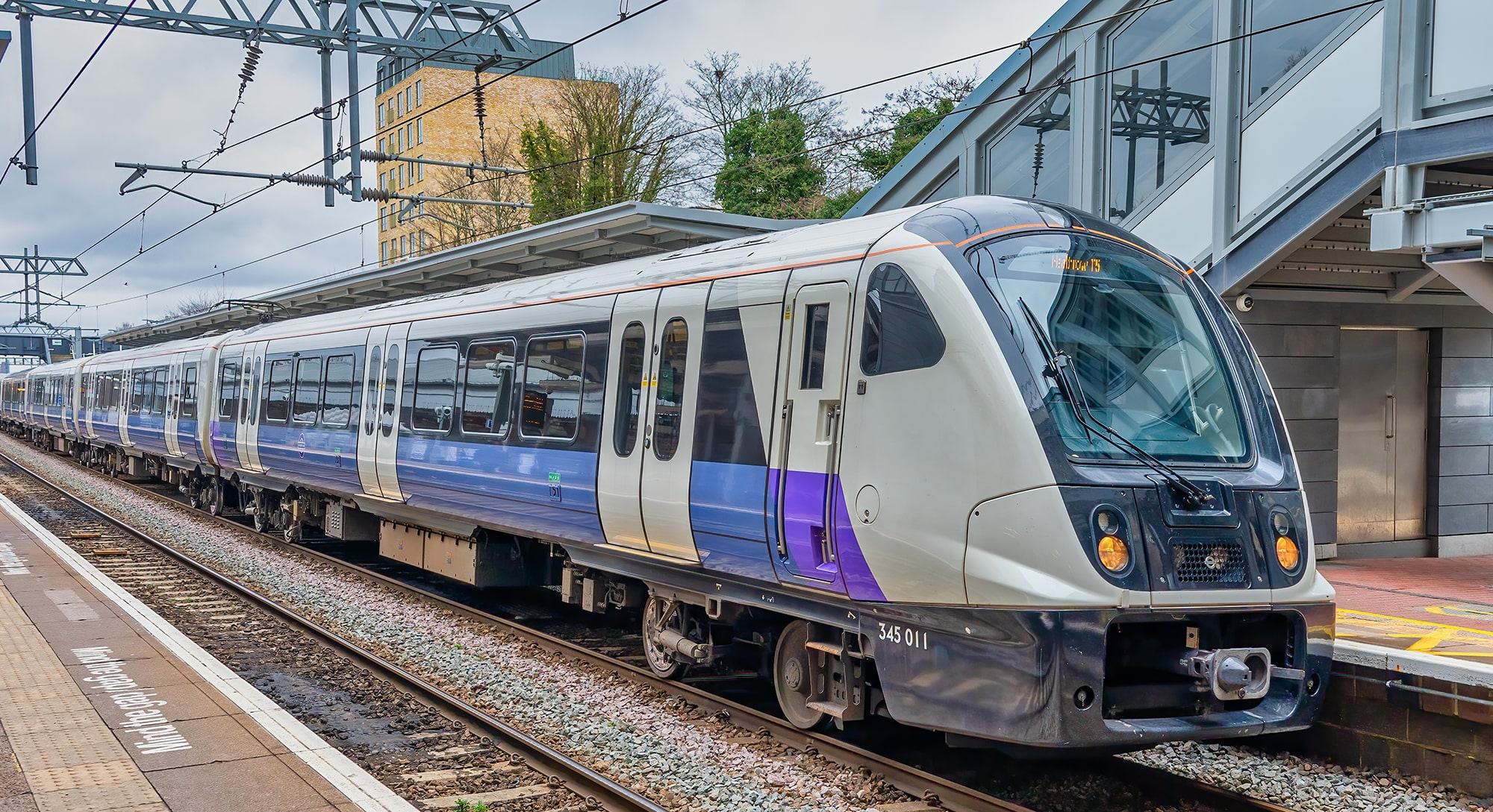 A modern passenger train painted white and purple waits at a station platform beneath overhead wires, with a footbridge and buildings in the background.