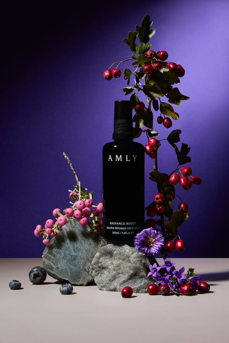 AMLY Radiance Boost Face Mist surrounded by flowers, berries and rocks on a dark purple background