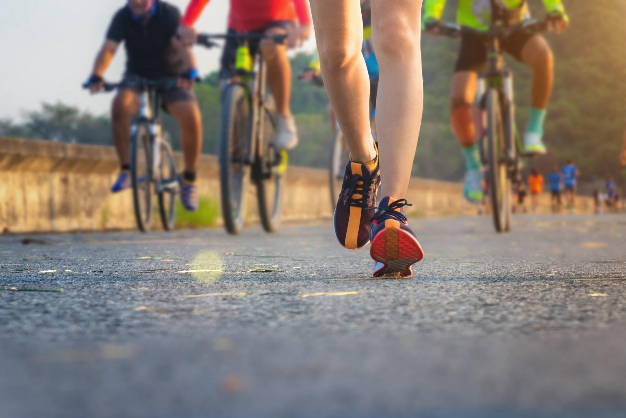 Close-up of a person walking or running on a paved road, with cyclists riding bikes in the background. The scene takes place outdoors, likely during a race or fitness event. The focus is on the persons legs and shoes.