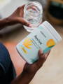 a woman's hands holding a glass of water in one and a jar of Brightcore's Revive collagen powder in the other.
