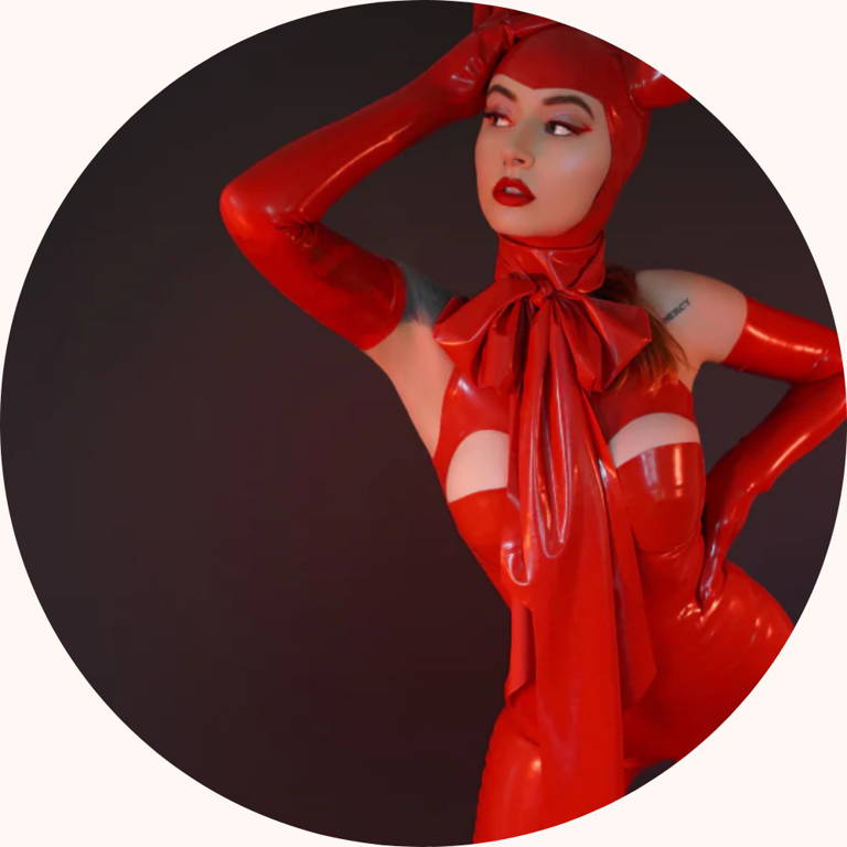 Model wearing Vex Clothing red latex Lucifer costume with cutout bodysuit, oversized bow, and matching hood.