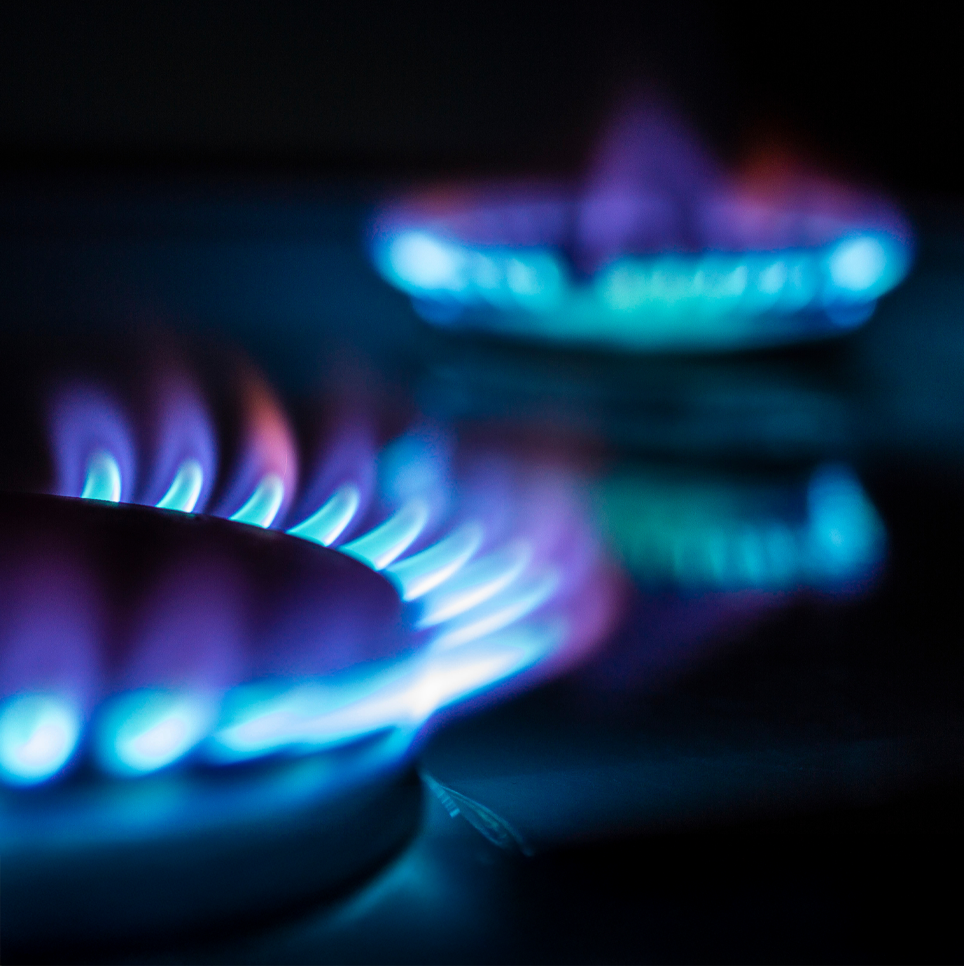 Close-up of blue flames on two gas stove burners, with one burner in sharp focus in the foreground and the other blurred in the background, set against a dark background.